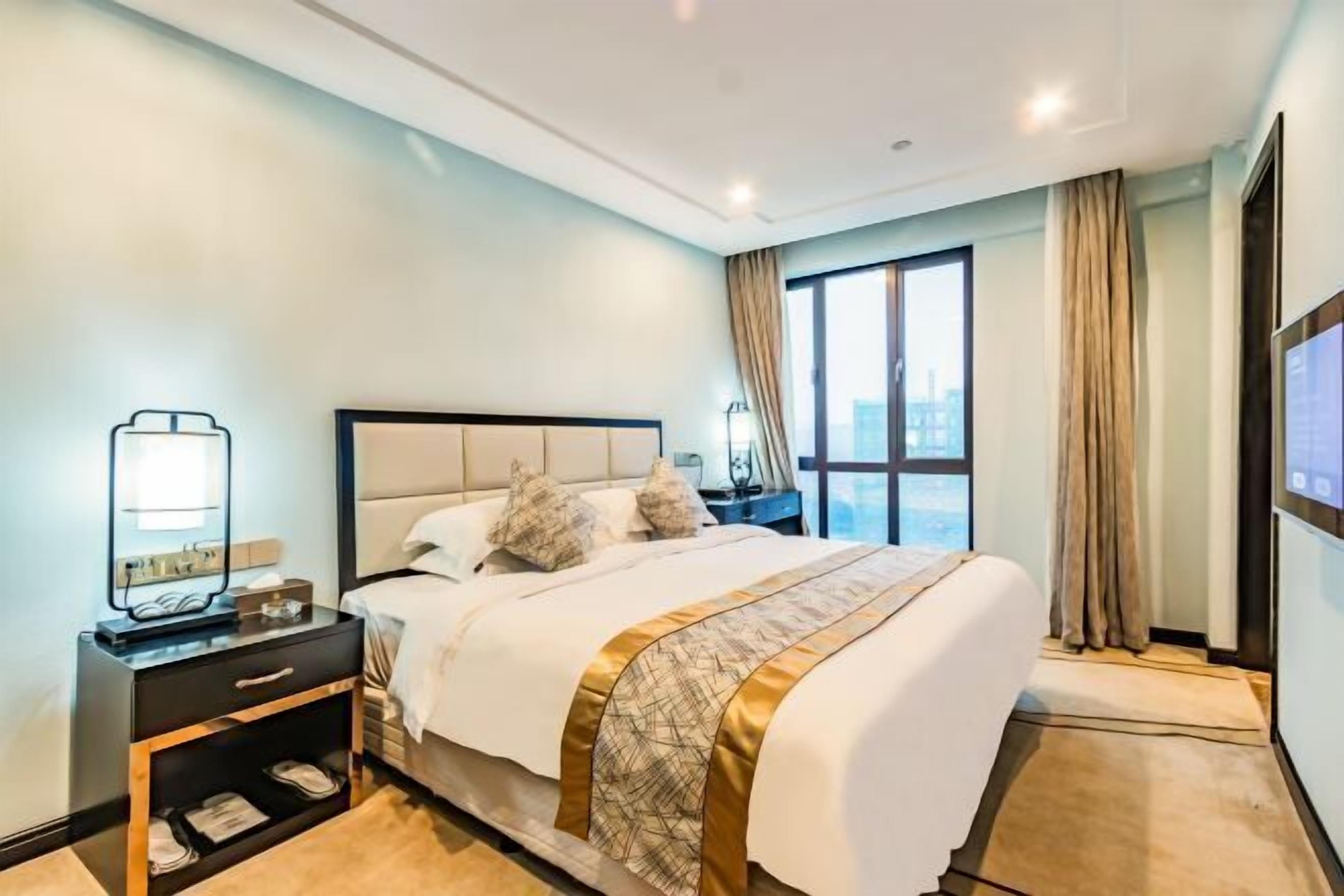 A hotel room with a large bed, a bedside table with a lamp, a TV, and a window with curtains.