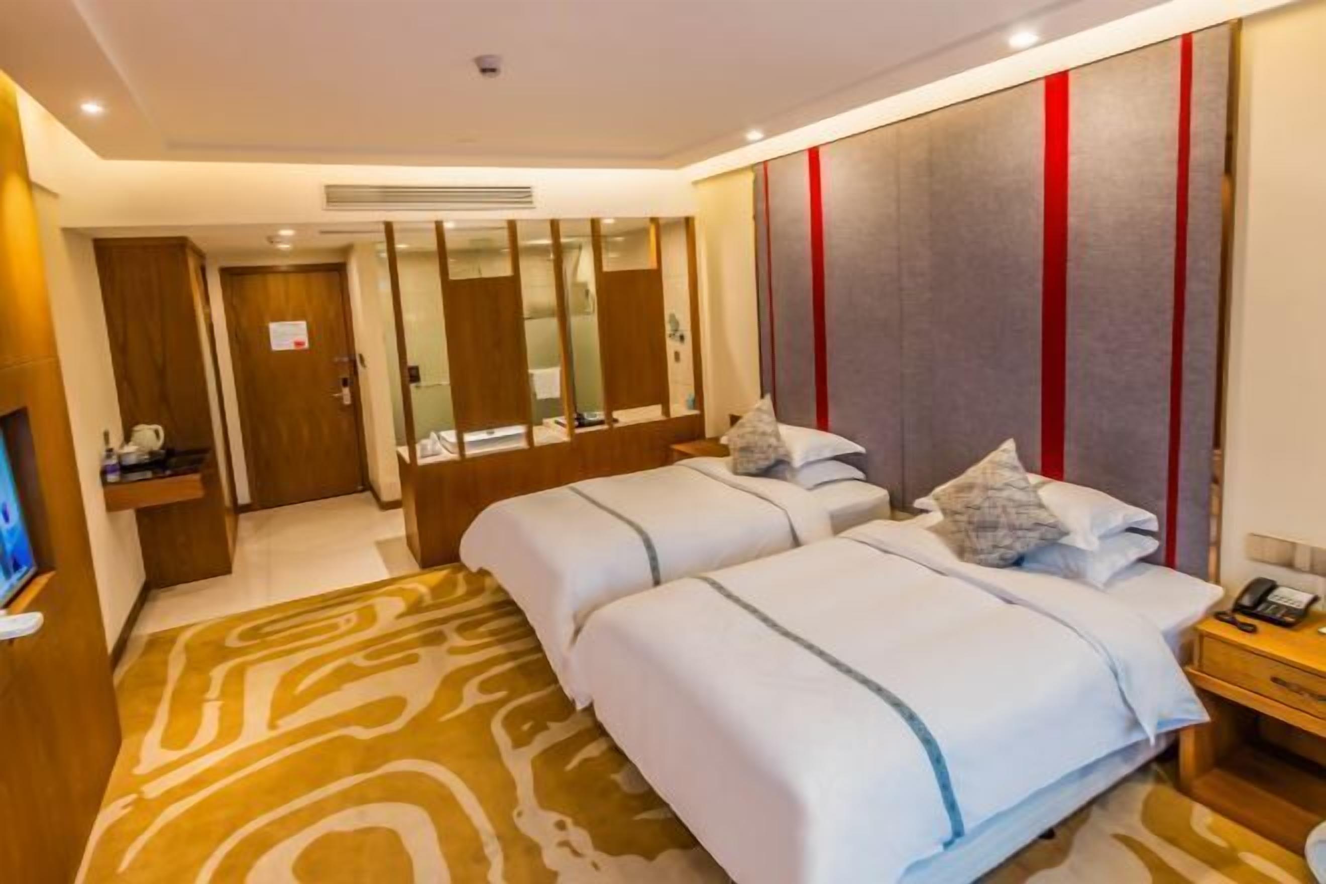 A hotel room with two beds, a TV, a desk, and a bathroom visible through a glass partition.