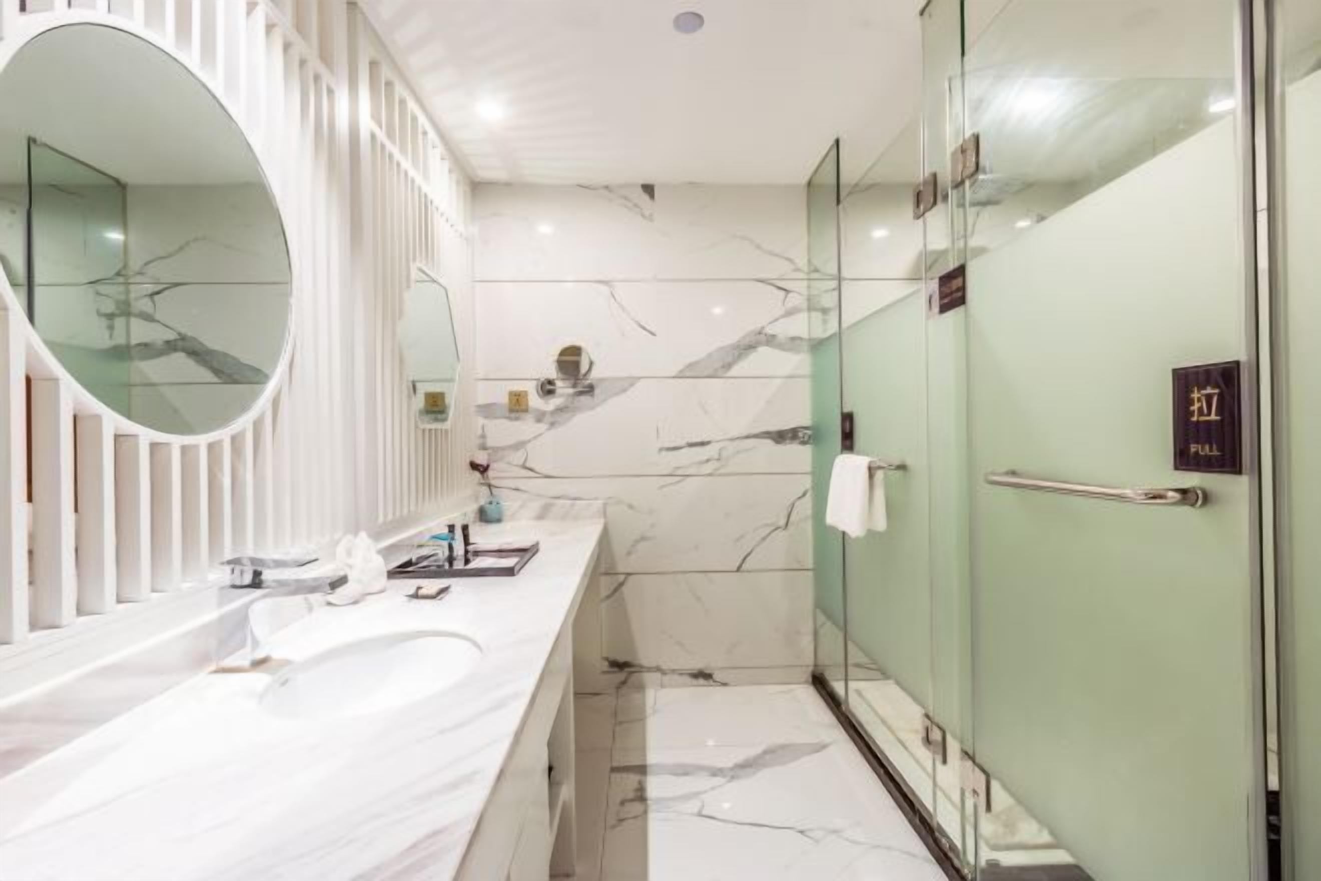 Un baño moderno con un espejo ovalado grande, un lavamanos de mármol blanco y una cabina de ducha de vidrio.