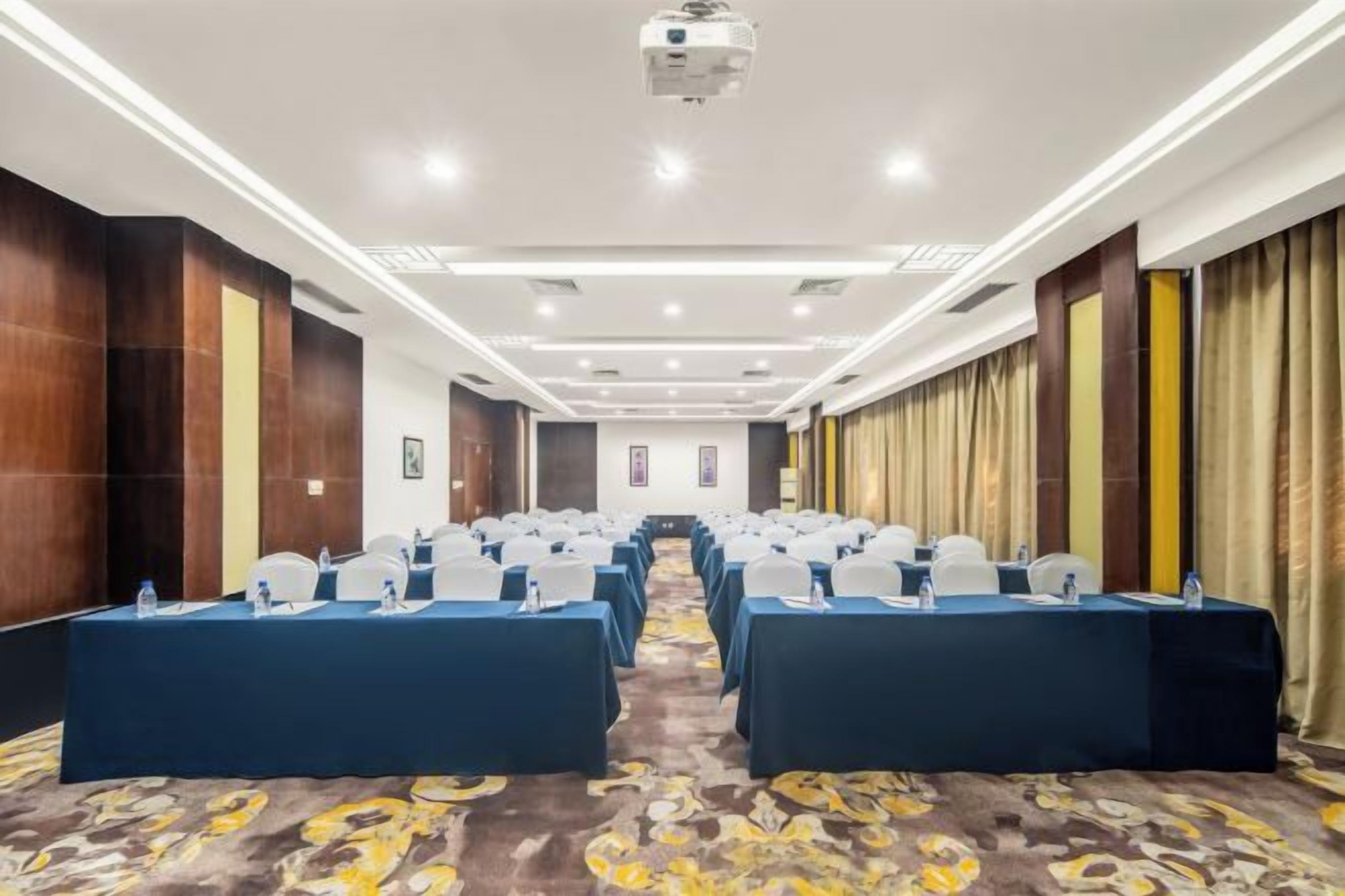 A conference room with rows of tables, each with a blue tablecloth and white chairs, equipped with microphones and water bottles.