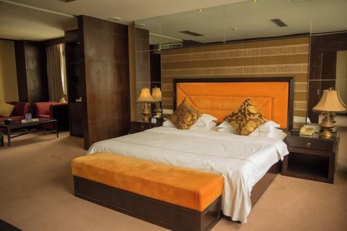 A hotel room with a large bed, a wooden headboard, two bedside tables with lamps, a small table with a chair, and a large mirror.