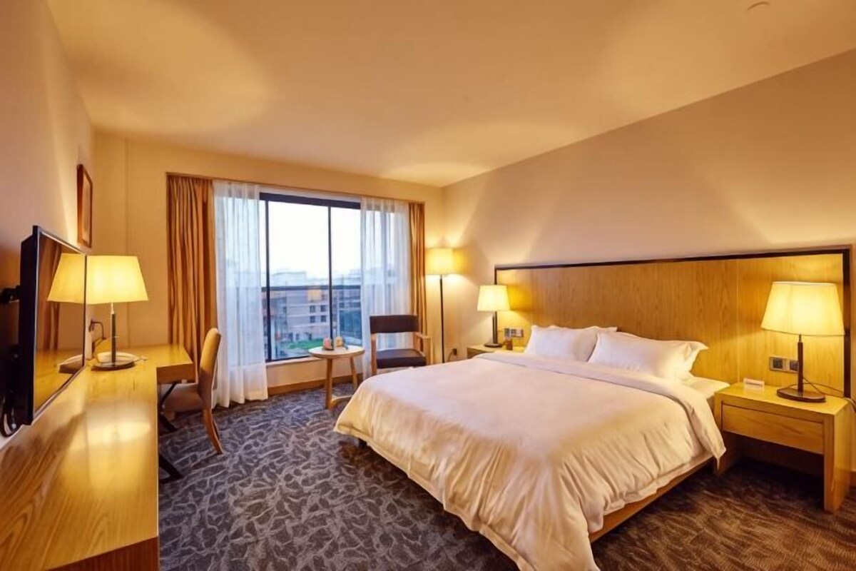 A hotel room with a large bed, two bedside tables with lamps, a desk with a chair, a large window with curtains, and a view of buildings.