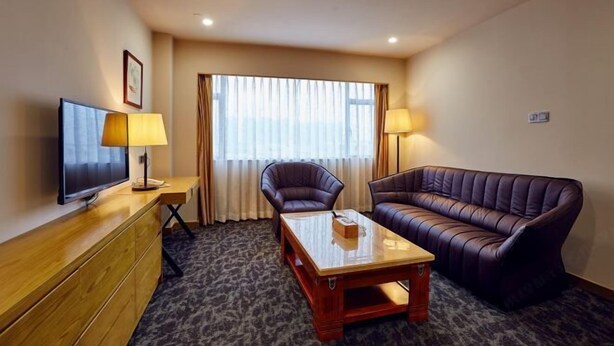 A hotel room with a television, a sofa, a chair, a table, and a window with curtains. - Xiya Hotel (Foshan)