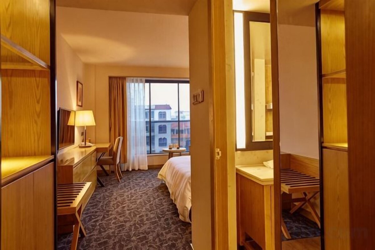 A hotel room with a bed, desk, chair, and a view of the city through large windows.