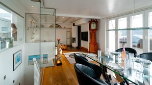 Ein modernes Wohnzimmer mit einem Esstisch aus Glas, gedeckt für sechs Personen, einer großen Holzuhr und Blick ins Freie durch große Fenster.
