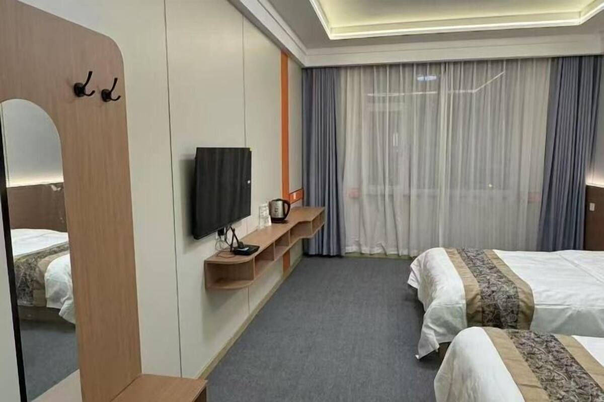 A hotel room with a large mirror, a flat-screen TV, a bed with a patterned bedspread, a bedside table, and a window with curtains.