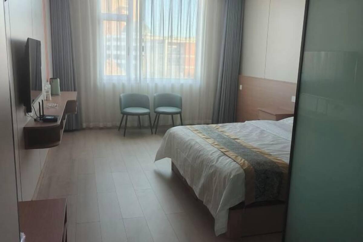 A hotel room with a bed, two chairs, a TV, and a window with curtains.
