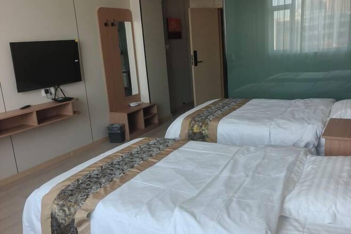 A hotel room with two beds, a television, a mirror, and a window with sheer curtains.
