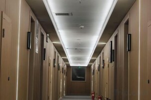Corredor de hotel com portas em ambos os lados, placas de saída de emergência e um extintor de incêndio à direita.