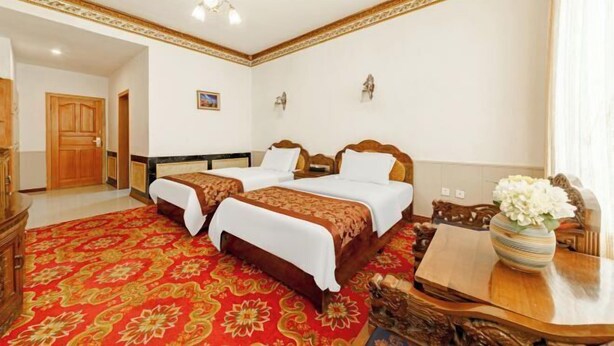 A hotel room with two beds, a wooden cabinet, a red patterned carpet, and a wooden table with a vase.