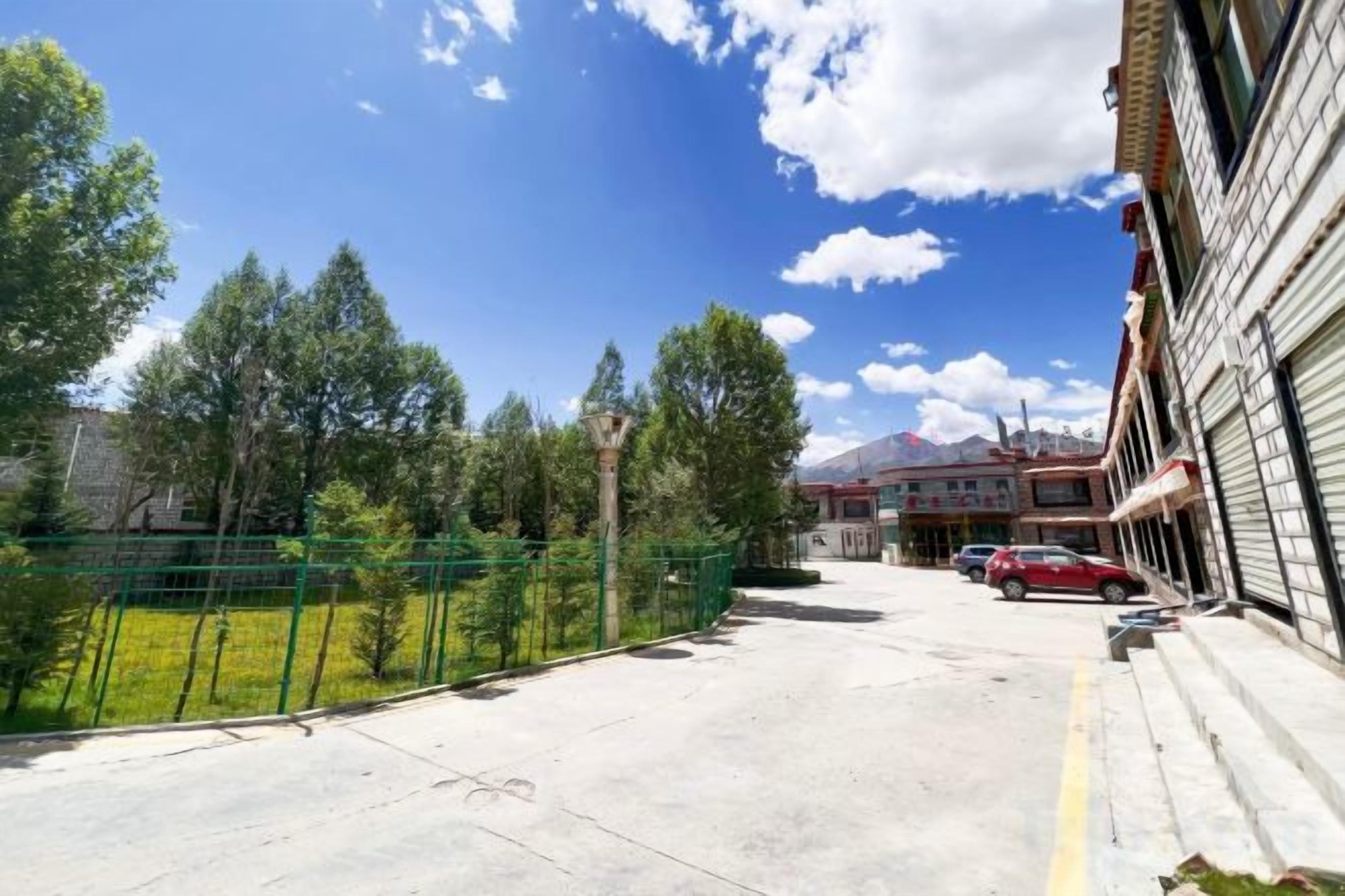 A paved road with a green fence, a building with a closed shutter, and a red car parked in the distance.
