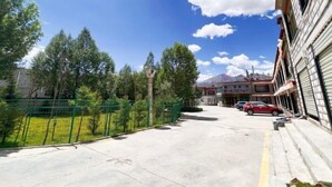 Vista de la calle con un edificio a la derecha, una cerca verde y árboles al fondo.
