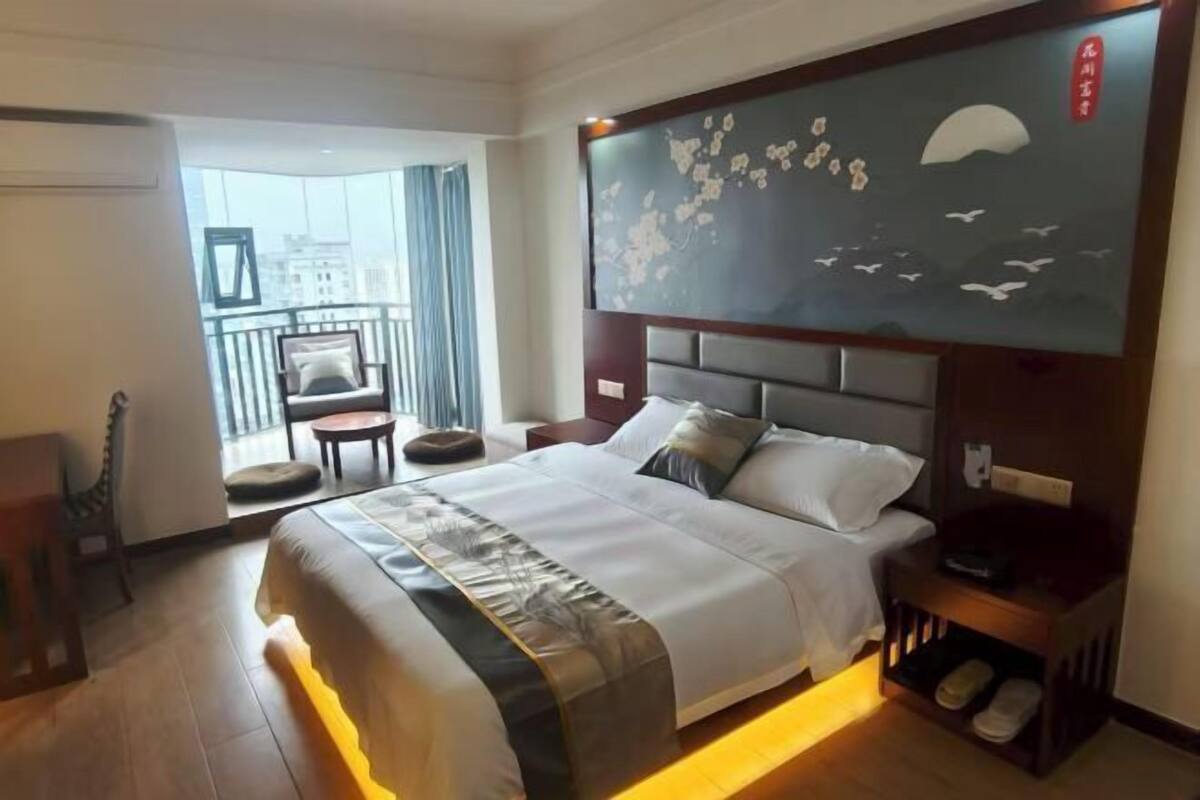 A hotel room with a bed, a nightstand, a chair, a balcony with a view, and a wall with a decorative painting.