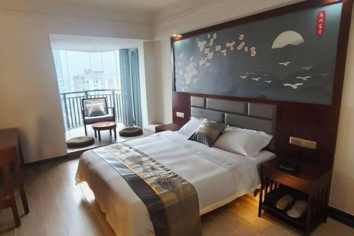 A hotel room with a large bed, a view of the city, a balcony with a chair, and a wall with a painting.