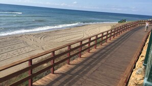 Un muelle de madera con barandilla metálica que conduce a una playa arenosa con surcos de neumáticos.