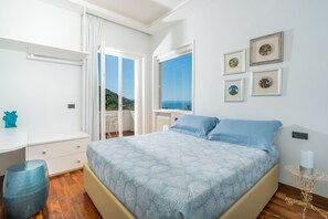 A bedroom with a bed, a desk, and a chair. There are windows with a view of the sea and hills. - MyExclusiveItaly - Luxury Villa in Sorrento Coast Swimming Pool & View (Vico Equense)