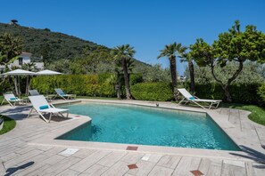 Une piscine rectangulaire, bordée de chaises longues et d’ombrelles, entourée de verdure et située au pied d’une colline.