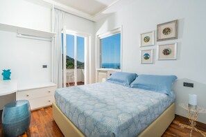 Une chambre à coucher avec un lit, un bureau et une chaise. Les fenêtres offrent une vue sur la mer et les collines.