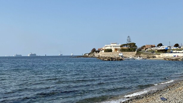 A coastal scene with a rocky shoreline, a clear blue sky, and several boats in the distance.