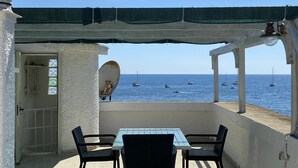 A covered outdoor seating area with a table and chairs, overlooking a sea with boats.