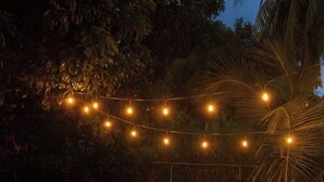 Ein Sitzbereich im Freien mit Holztisch und -sofa, umgeben von Lichterketten und einem kleinen Feuerkorb.