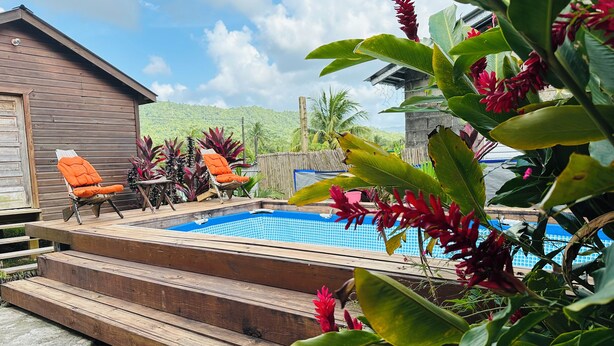 Eine Holzterrasse mit Pool, umgeben von tropischen Pflanzen und zwei orangefarbenen Liegestühlen.