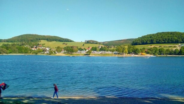 A lake with a small town in the background, a person walking on the shore, and a boat in the water. - Neles Kate am Diemelsee by Interhome (Marsberg)