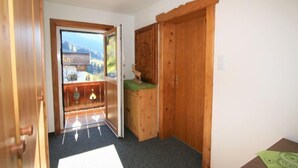 Ein Zimmer mit Holztür, die auf einen Balkon mit Bergblick führt, einem kleinen Holzkasten und einer grünen Matte auf dem Boden.