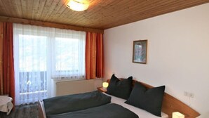Ein Schlafzimmer mit Holzbalkendecke, einem Bett mit dunkler Bettwäsche, einem Nachttisch mit Lampe und einem Fenster mit Vorhängen.