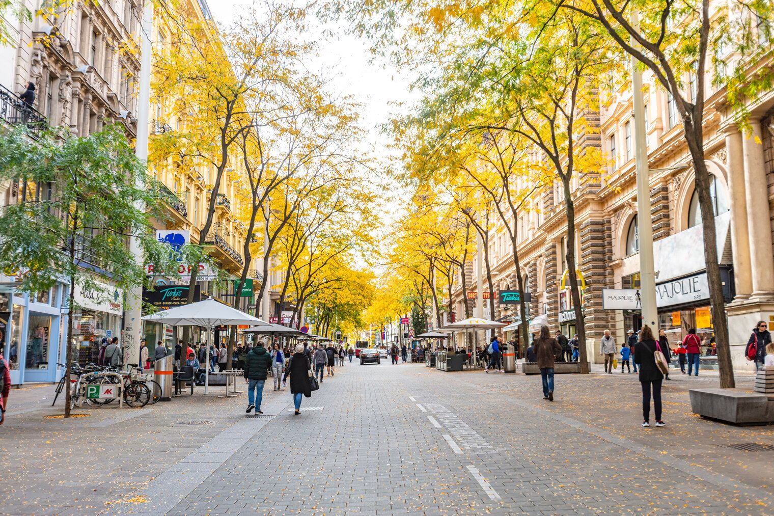 A bustling urban street lined with trees in autumn foliage, shops, and cafes.