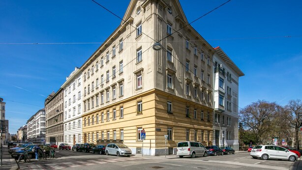 Ein großes, historisches Gebäude mit klassischer Architektur, das mehrere Stockwerke und einen markanten Hauseingang im Winkel aufweist.