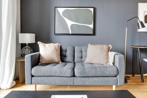 A grey sofa with two beige pillows, a framed abstract painting, a floor lamp, and a small table with a lamp.