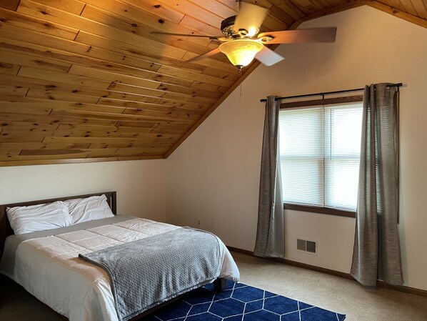 A bedroom with a sloped ceiling, a bed with a gray blanket, a ceiling fan, and a window with curtains. - East River Retreat, WATERFRONT+ Fire Pit+Pet friendly (Two Rivers)