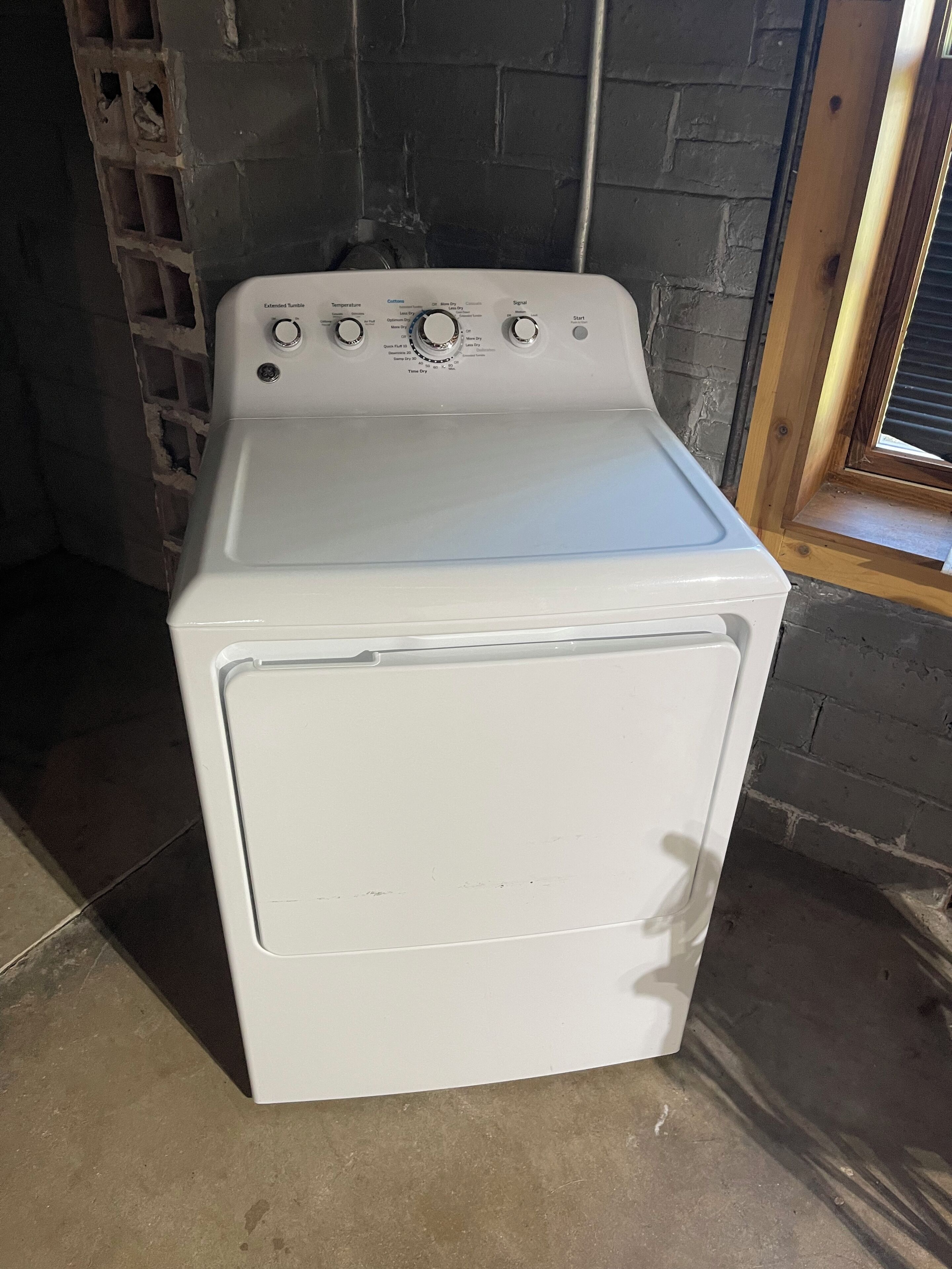 Una secadora eléctrica blanca en una habitación con pared de ladrillo y ventana.