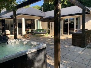 A hot tub in a covered outdoor area with a patio, a wooden bench, and a view of a house in the background.