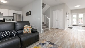 A modern living room with a black leather sofa, a patterned throw pillow, and a wooden floor. A staircase leads to a second floor. - Luxury Entire house mins Niagara falls & Brock uni (Thorold)