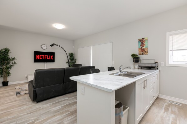 A modern living space with a kitchen island, a black sofa, a white countertop, and a wall-mounted TV displaying "NETFLIX".