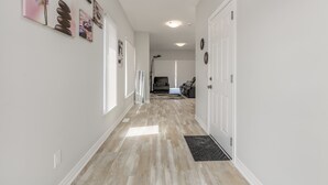 A hallway with a light wood floor, white walls, and a door at the end leading to a living area with a couch and a rug. - Luxury Entire house mins Niagara falls & Brock uni (Thorold)
