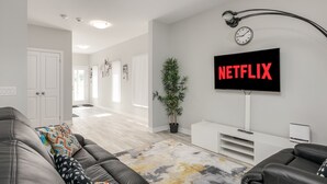 A modern living room with a large flat-screen TV mounted on the wall, a potted plant, and a grey sofa with patterned cushions. - Luxury Entire house mins Niagara falls & Brock uni (Thorold)