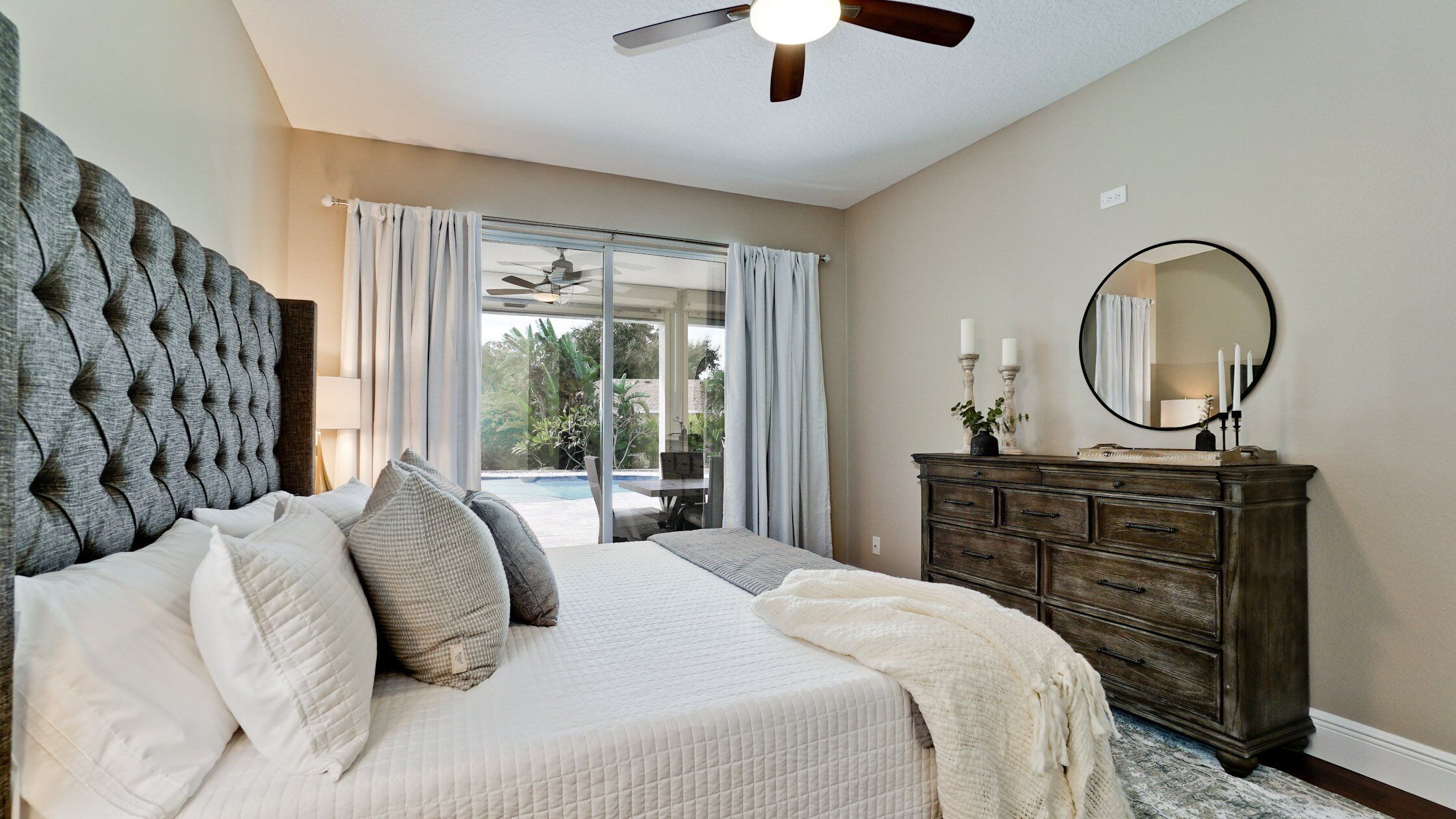 A bedroom with a large bed, a dresser, a ceiling fan, and a view of a pool.