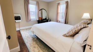 A bedroom with a bed, two windows with curtains, a bedside lamp, a dresser, a mirror, and a radiator. - New River Cottage (Crumpler)