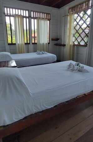 A room with two beds, each with white linens and towels, wooden headboards, and windows with curtains.