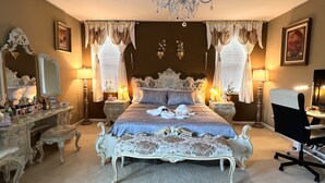 Ein klassisches Schlafzimmer mit einem großen, verzierten Bett, zwei Nachttischen mit Lampen, einem Schreibtisch mit Stuhl und einem Kronleuchter.
