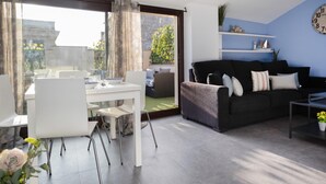 A modern living room with a black sofa, white dining table, and a large sliding glass door leading to a balcony.