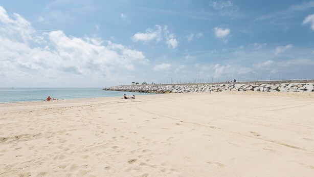 A wide sandy beach with footprints, a rocky breakwater, and a clear blue sky.