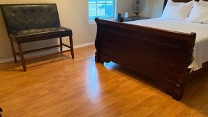 Une chambre avec un plancher en bois, un banc en cuir noir, un cadre de lit en bois et une couvre-lit blanche.