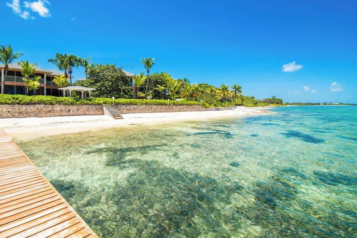 Una vista chiara di un resort sulla spiaggia, con un pontile di legno che conduce alla riva, palme e cielo azzurro.