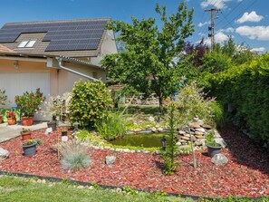 A residential garden with a pond, solar panels on the roof, and various potted plants.
