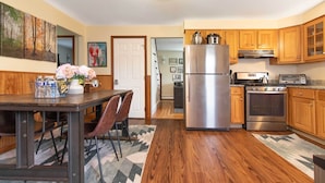 A kitchen with wooden cabinets, a stainless steel refrigerator, a stove, and a dining area with a table and chairs. - Pet-Friendly Cranston Home w/ Fire Pit & BBQ! (Cranston)