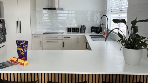 Een moderne keuken met witte kasten, een centraal kookeiland met barstoelen, een inbouwoven en een raam met jaloezieën.